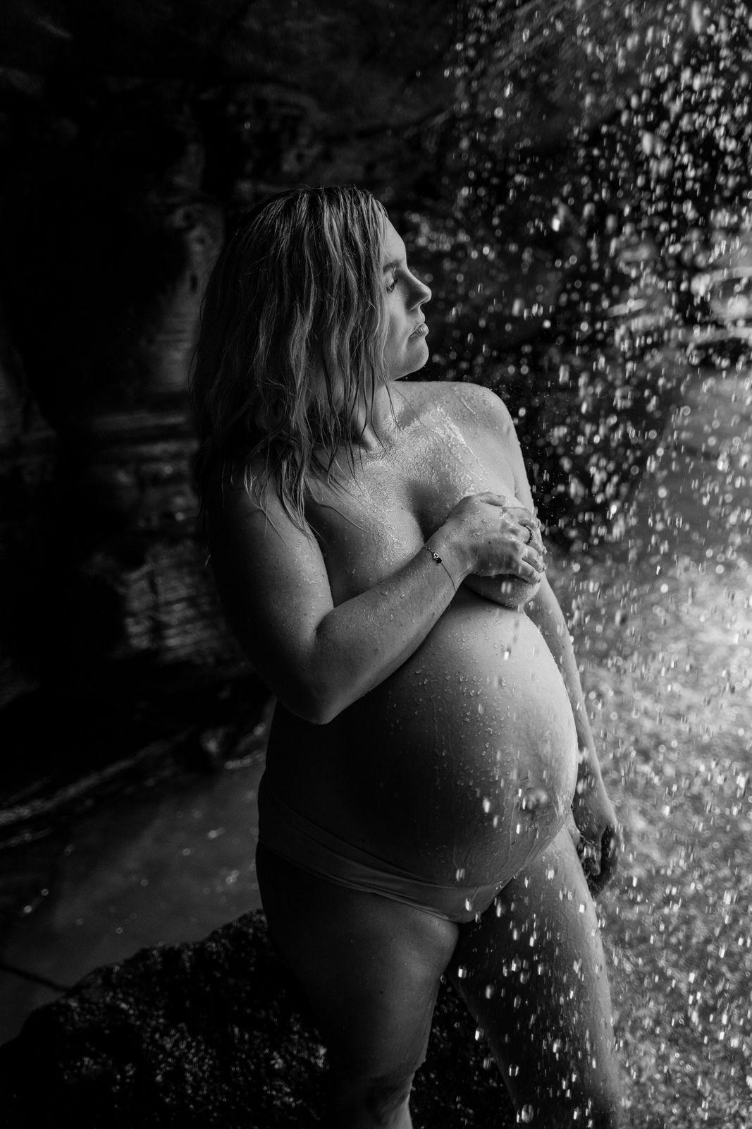 Black and white maternity portrait of a mum-to-be cradling her bump beside a waterfall