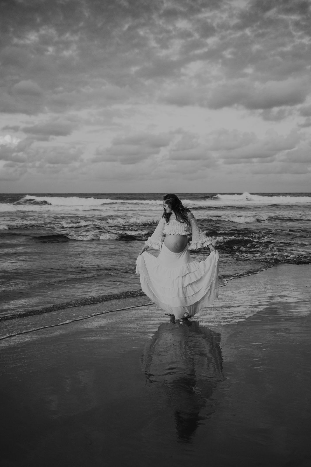 Black and white photo of a mum-to-be in a flowing white dress standing in the shallows