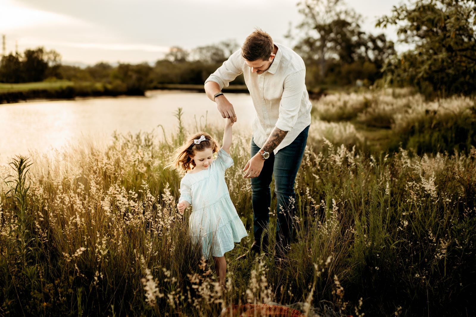 Dad holding his little girl's hand as she walks through long golden grass by the river