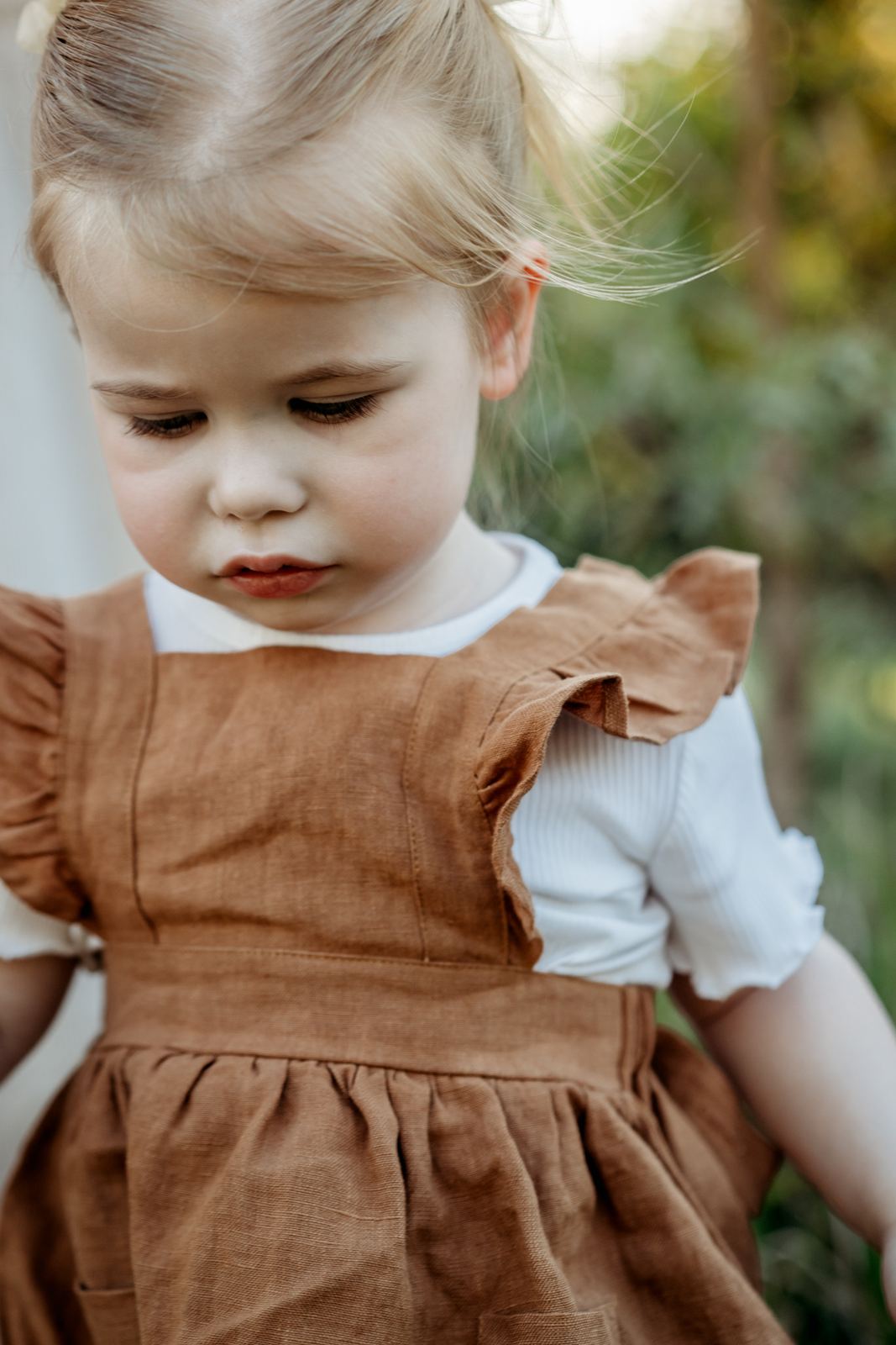 Little girl in a rust linen pinafore looking down, soft green trees behind her