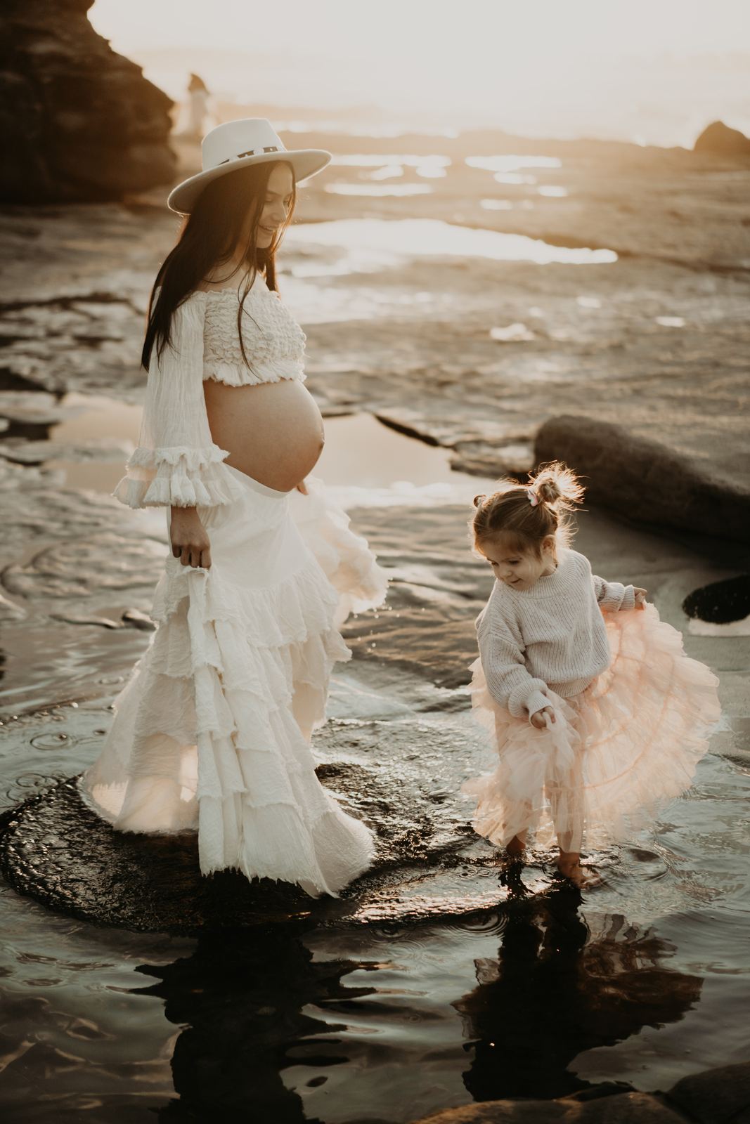 Pregnant mum in a white dress walking through a rock pool with her toddler