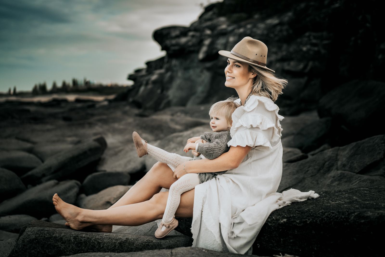 Mum in a sun hat sitting on dark rocks with her toddler on her lap
