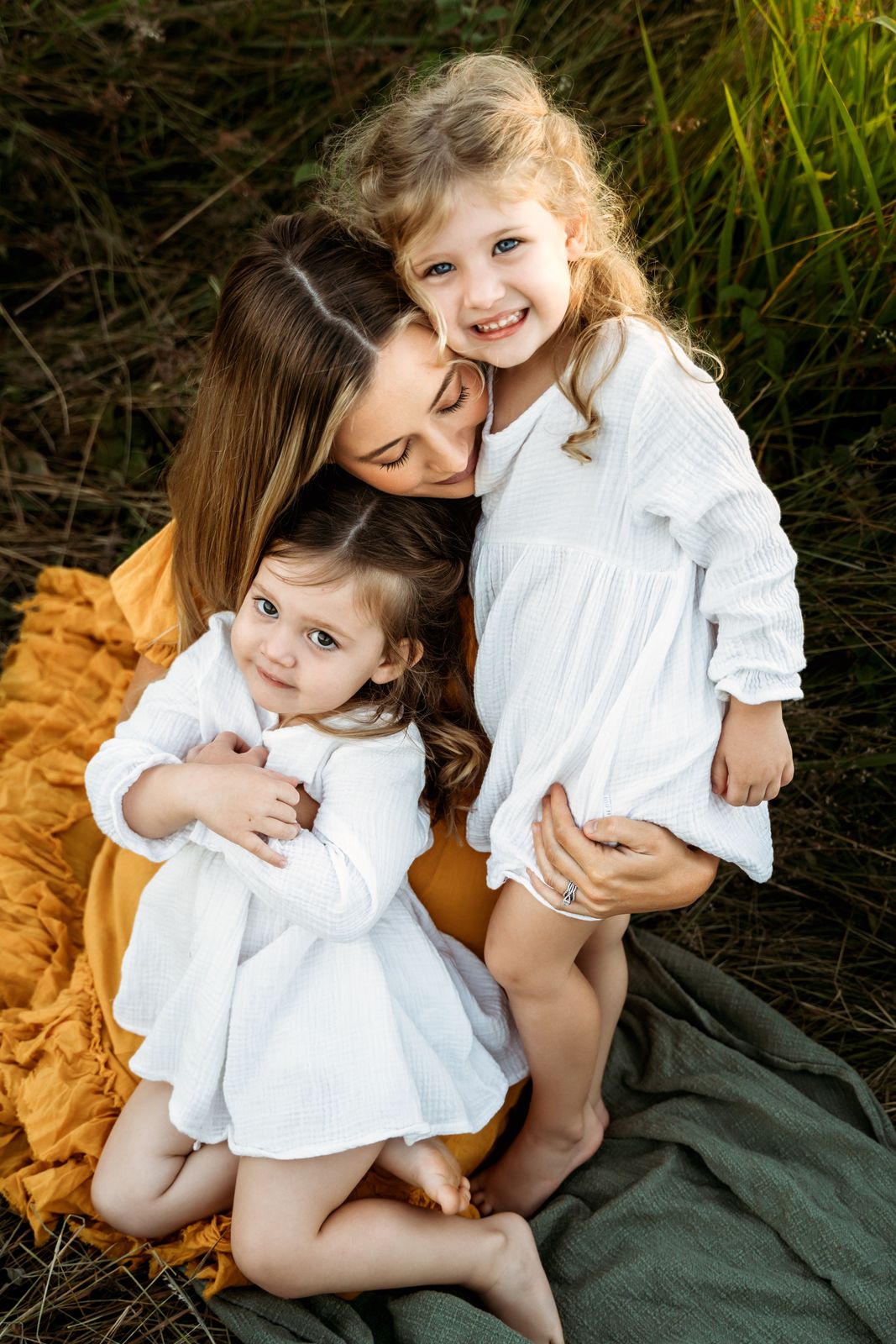 Mum hugging her two little girls in white dresses in long grass