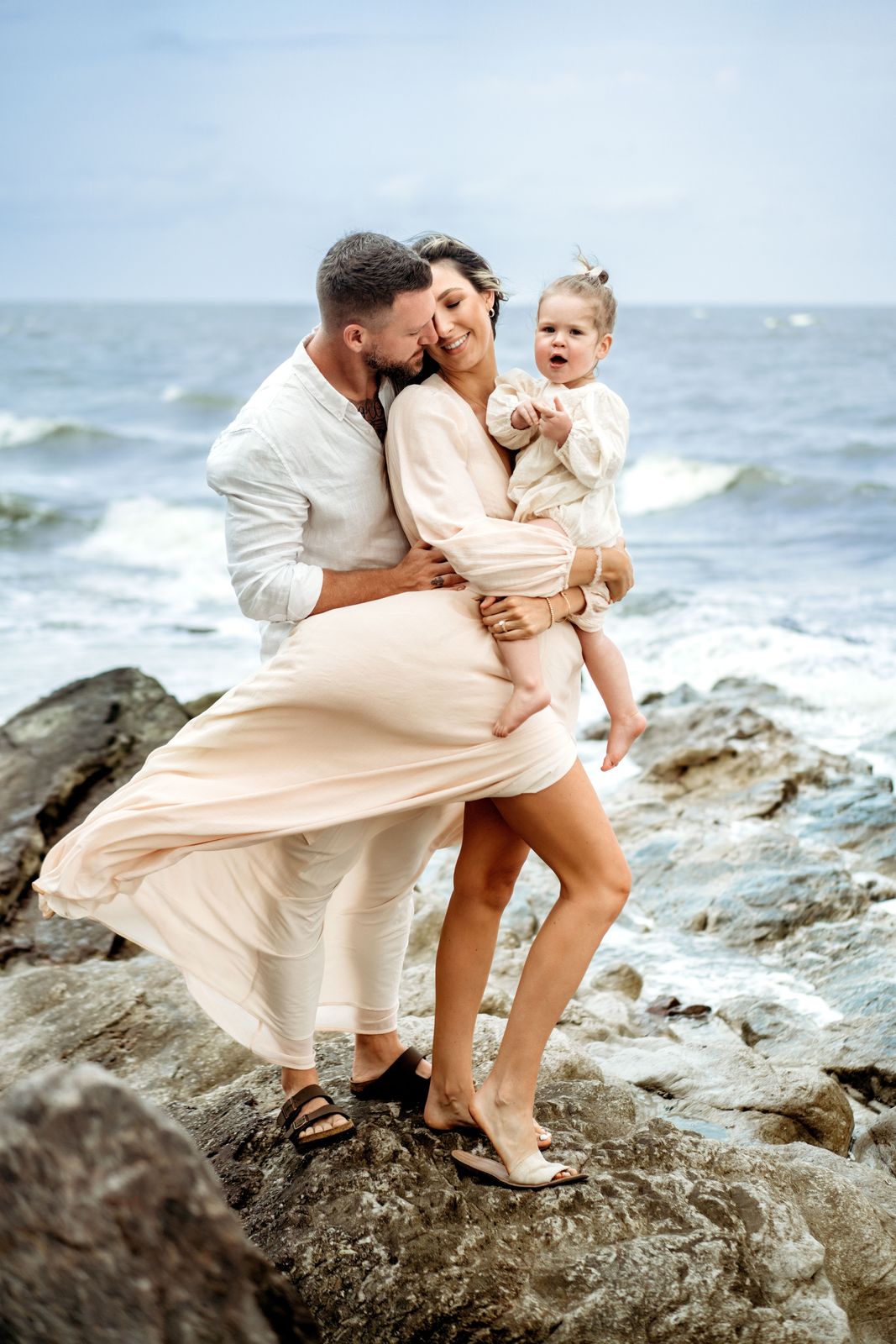 Parents and their toddler on the rocks with the sea wind in her dress