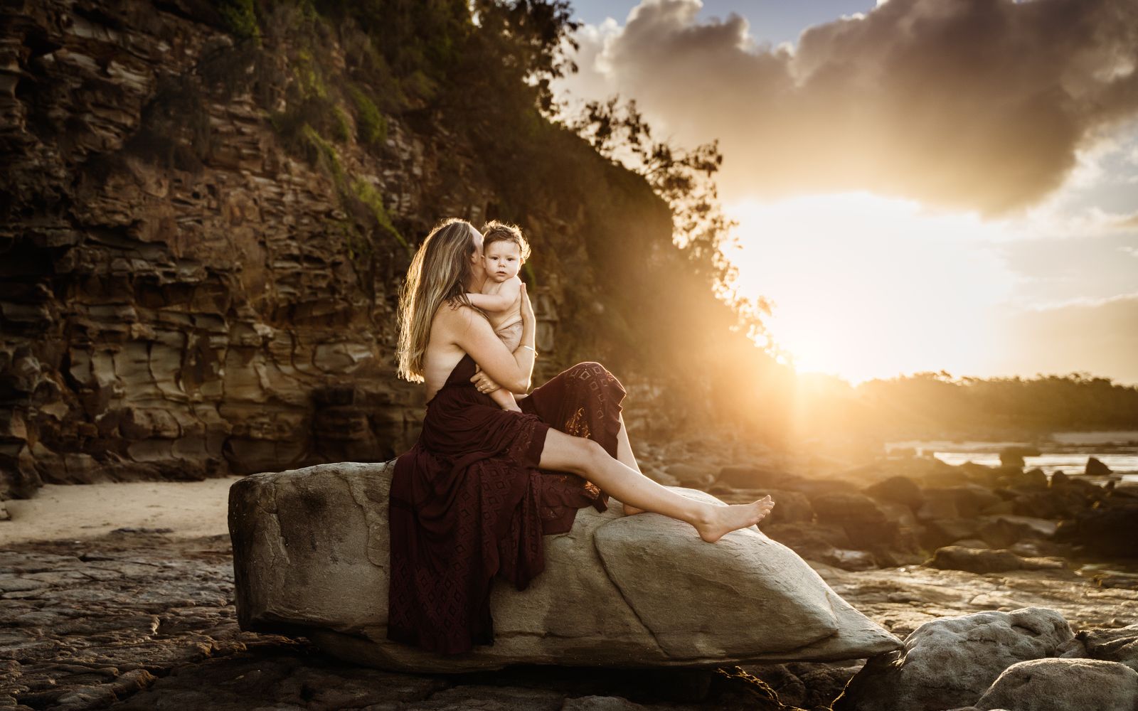 Mum in a red dress sitting on a sandstone rock as the sun sets behind her